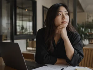 An Asian woman sitting thoughtfully beside a laptop and workspace notes, representing UX strategy and content marketing planning in a calm, professional office setting.