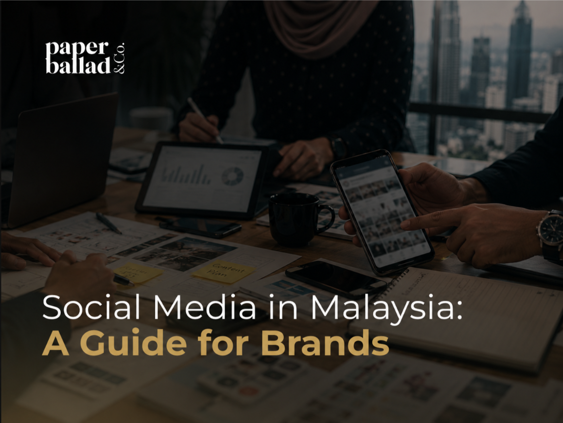 A dark, professional cover image showing a team discussing digital content strategy around a meeting table, with a phone, tablet, laptop, printed layouts, notes and blurred analytics visuals. The background shows a modern city skyline, giving the image a business and marketing feel. The Paperballad & Co logo appears at the top left, with the headline “Social Media in Malaysia: A Guide for Brands” displayed in large white and gold text across the lower section.