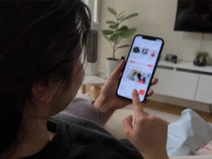 A woman sits at home scrolling through a shopping app on her phone, with the screen intentionally blurred and no clear product details visible.