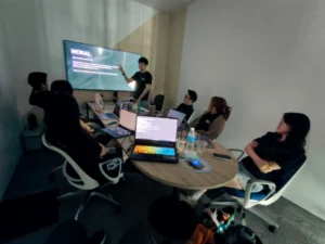 A team gathers in a meeting room while a colleague presents on a large screen, with laptops, notebooks, and drinks spread across the table as everyone discusses strategy and content planning. The collaborative office scene reflects how businesses approach social media in Malaysia through teamwork, research, and campaign planning.