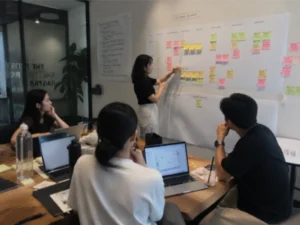 A team collaborates in a workshop around laptops, sticky notes, and a customer journey map while discussing ideas and planning solutions together, illustrating how UX strategy in Malaysia begins with research, collaboration, and understanding customer experiences.