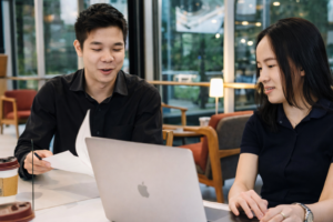 Two colleagues collaborate over a laptop in a modern café setting, discussing ideas and reviewing documents - illustrating what a UX strategist does by planning user-focused digital experiences and workflows.