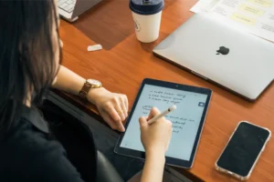 A person writes notes on a tablet at a desk with a laptop and coffee nearby, illustrating what a UX strategist does-planning user experiences, defining strategy, and organizing ideas for digital products.