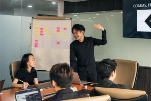 A team discussion in a meeting room, with one person presenting beside a strategy board while colleagues listen and take notes around the table discussing what is a brand.