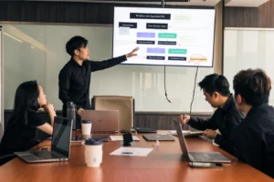 A team meeting in progress about what is a brand, with one person presenting a workflow on screen while colleagues discuss, take notes, and review laptops around the conference table.