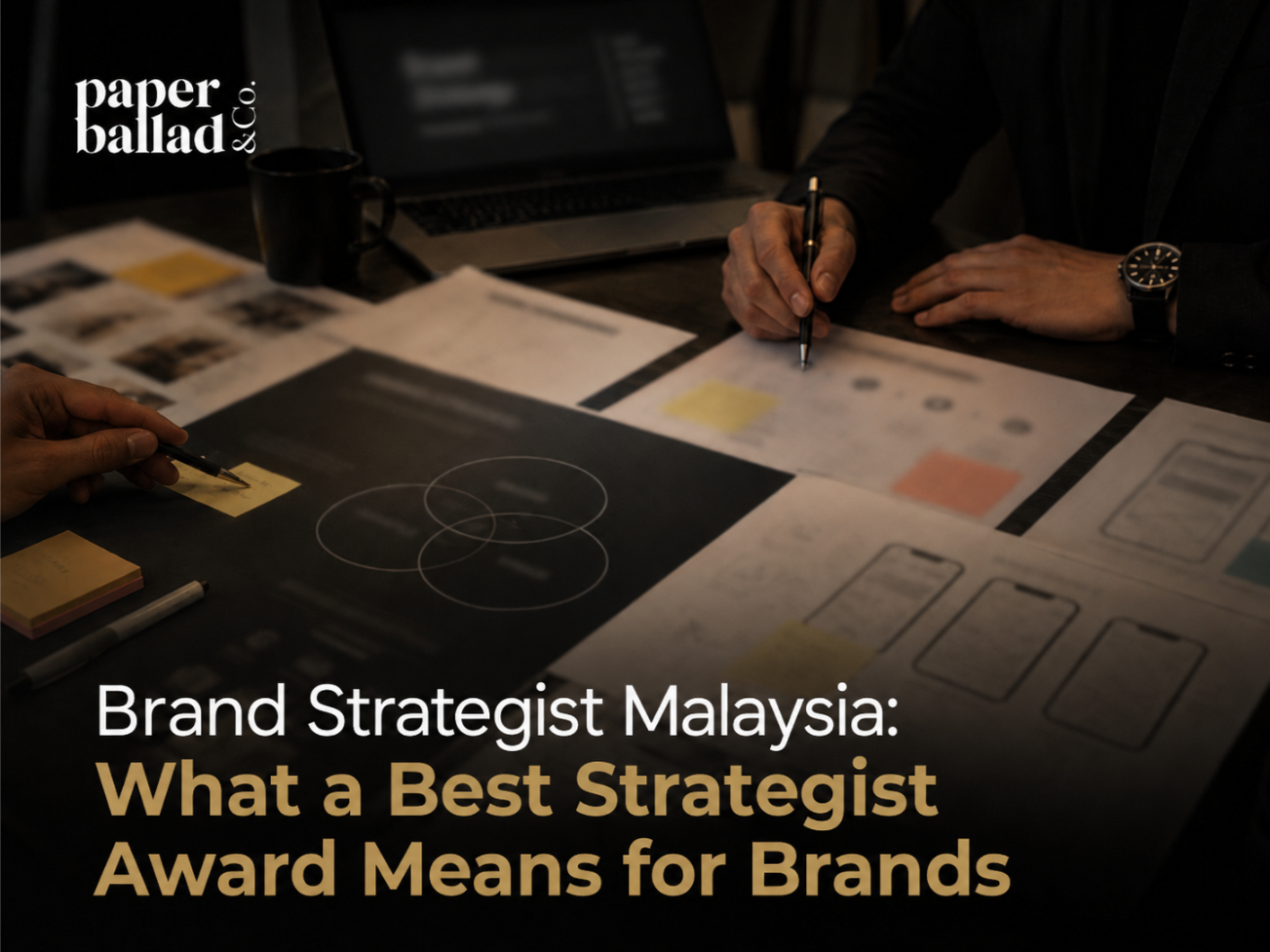 A dark, premium cover image showing a Brand Strategist workspace with hands reviewing brand strategy documents, audience mapping, positioning notes, wireframes and sticky notes on a table. The background text on the documents and laptop is softly blurred, while the Paperballad & Co logo appears clearly at the top left. The headline “Brand Strategist Malaysia: What a Best Strategist Award Means for Brands” is displayed in large white and gold text across the lower section.