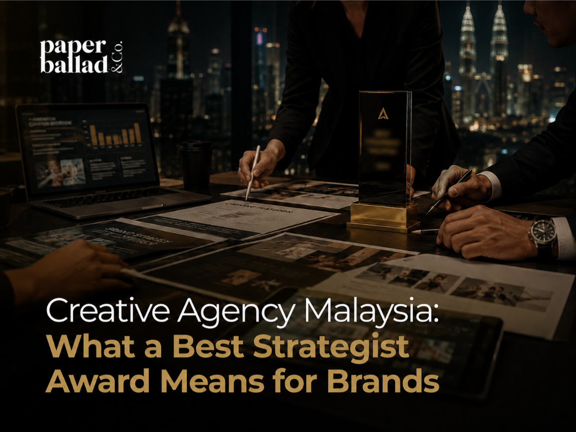 A dark, premium cover image showing a creative agency strategy meeting in a modern office at night, with a blurred Kuala Lumpur skyline in the background. A team reviews brand strategy documents, campaign layouts and analytics on a laptop while an award trophy sits on the table with its wording softly blurred. The Paperballad & Co logo appears clearly at the top left, with the headline “Creative Agency Malaysia: What a Best Strategist Award Means for Brands” displayed in large white and gold text across the lower section.