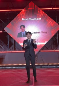 Creative Agency Malaysia Blog Post - Paperballad CEO Casimir Yong stands in a black tuxedo stands on stage holding an award while smiling and giving a thumbs up, with a large screen behind him announcing his Best Strategist recognition. The image highlights a creative agency in Malaysia celebrating an industry achievement for strategic excellence.
