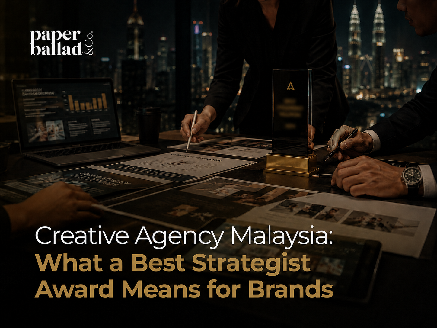 A dark, premium cover image showing a creative agency strategy meeting in a modern office at night, with a blurred Kuala Lumpur skyline in the background. A team reviews campaign documents, brand strategy layouts and analytics on a laptop while an award trophy sits on the table. The Paperballad & Co logo appears at the top left, with the headline “Creative Agency Malaysia: What a Best Strategist Award Means for Brands” displayed in large white and gold text across the lower section.