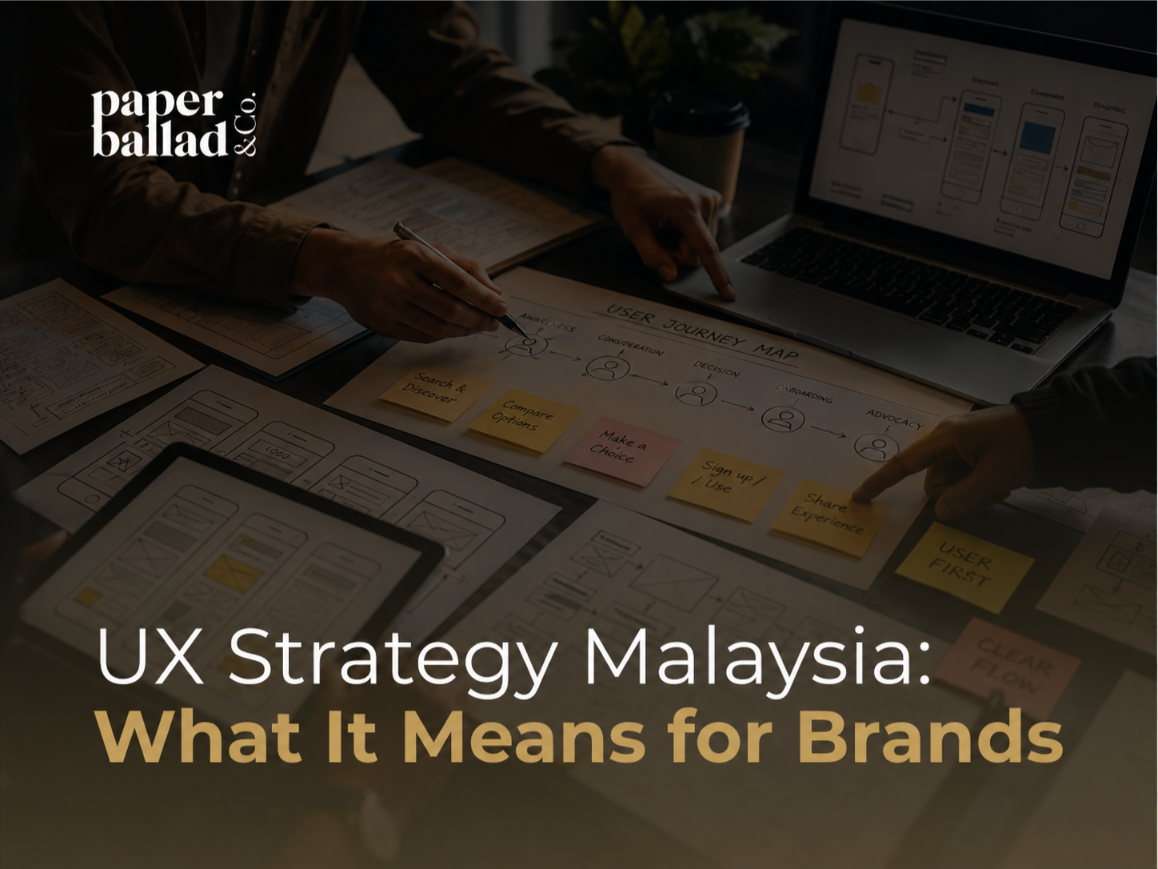 A dark, premium cover image showing a UX strategy workspace with hands reviewing user journey maps, wireframes, sticky notes and digital interface sketches on a table. A laptop displays mobile app flow diagrams while the Paperballad & Co logo appears clearly at the top left. The headline “UX Strategy Malaysia: What It Means for Brands” is displayed in large white and gold text across the lower section.