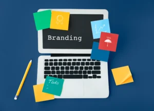 brand awareness illustrated through a top-down view of a laptop with the word “Branding” on its screen. Colourful sticky notes surround the laptop, showing icons like a medal, a graph, and an umbrella, along with a “Todo” note. A pencil lies beside it. The scene represents planning, strategy, and creative work involved in building a memorable brand.